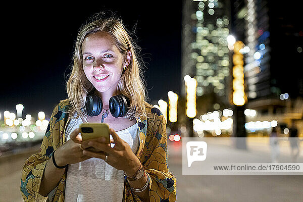 Fröhliche junge blonde Frau mit kabellosen Kopfhörern  die nachts ihr Mobiltelefon in der Hand hält