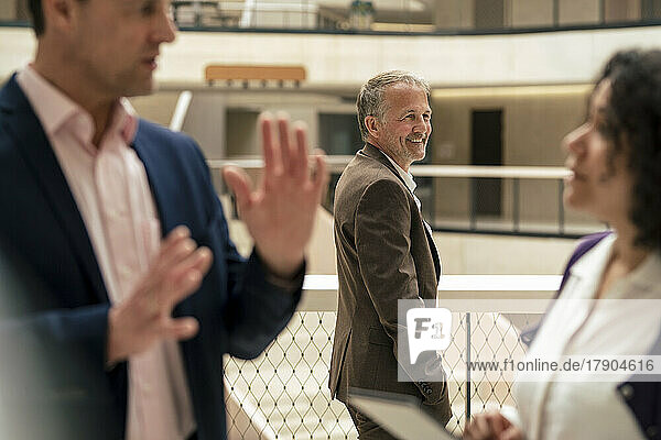 Smiling businessman standing by railing with colleagues discussing in foreground at office