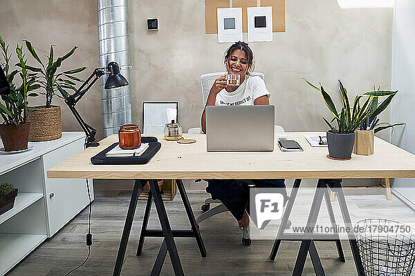 Smiling businesswoman drinking tea doing video call through laptop at home office
