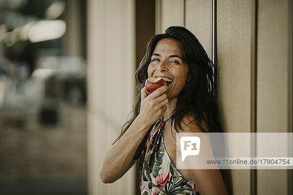 Happy mature woman eating apple by wall