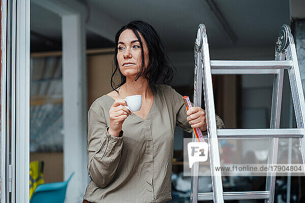 Nachdenkliche Frau hält Kaffeetasse in der Hand und steht zu Hause an der Leiter