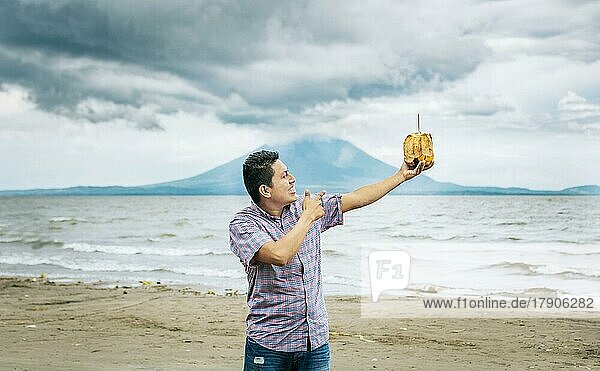 Junger Mann im Urlaub und genießt eine Kokosnuss am Strand. Lächelnder Mann  der eine Kokosnuss an einem Strand in Nicaragua hält und auf sie zeigt. Junger Mann  der auf eine Kokosnuss am Strand zeigt