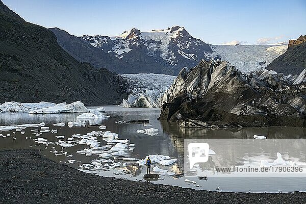 Wanderer steht auf einem Felsen im See  Spiegelung in der Gletscherlagune Svínafellslon mit Eisschollen  Gletscherzunge Svínasfellsjökull  hinten vergletscherter Berggipfel Hrútfjallstindar  bei Sonnenuntergang  Svínafell  Island  Europa