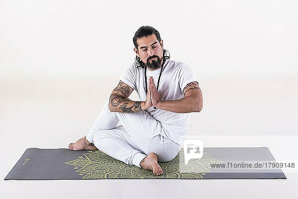 Ein weiß gekleideter Mann macht Yoga auf einer Matte vor weißem Hintergrund. Vorderansicht