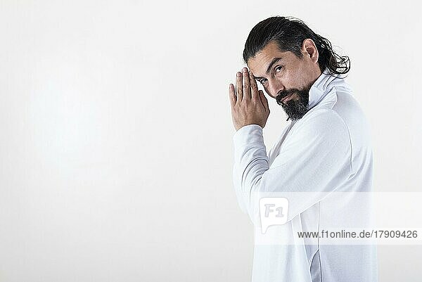 A man dressed in white clasping hands while looking at camera over white background. Tadasana yoga pose. Copy space. Studio shot