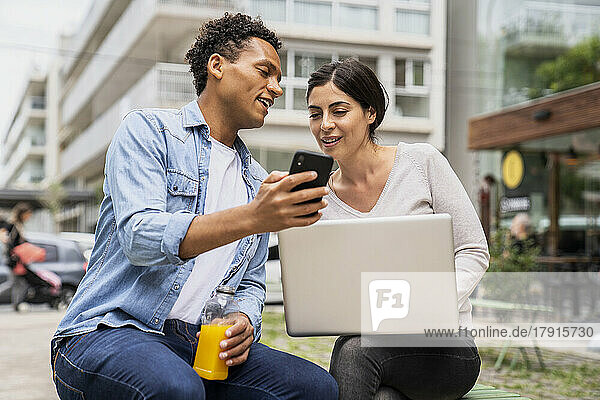Front view shot of diverse couple hanging out and having fun with their smartphones and computer in an outdoor setting