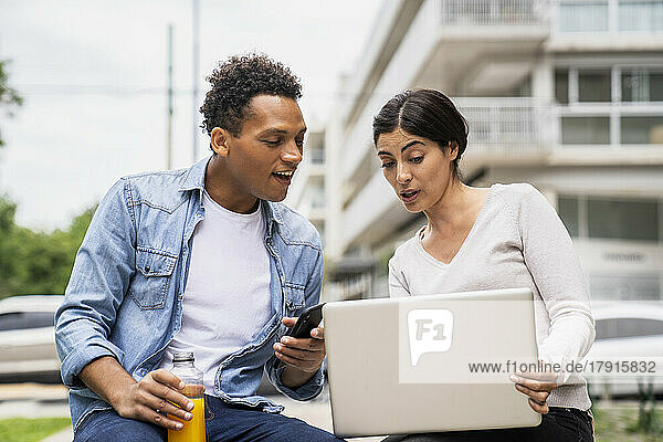 Mittelaufnahme eines afroamerikanischen Mannes und einer lateinamerikanischen Frau  die bei der Arbeit im Freien einen Laptop benutzen