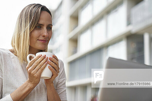 Vorderansicht einer Frau  die eine Tasse Kaffee in der Hand hält und auf den Bildschirm eines Laptops blickt