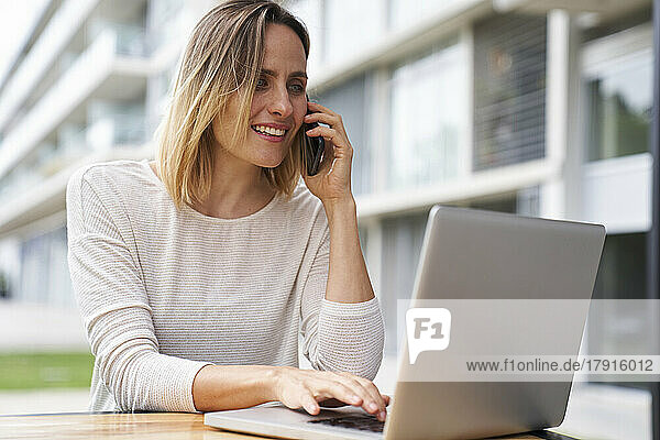 Mid-shot portrait of woman working outdoors on laptop computer while speaking on mobile phone