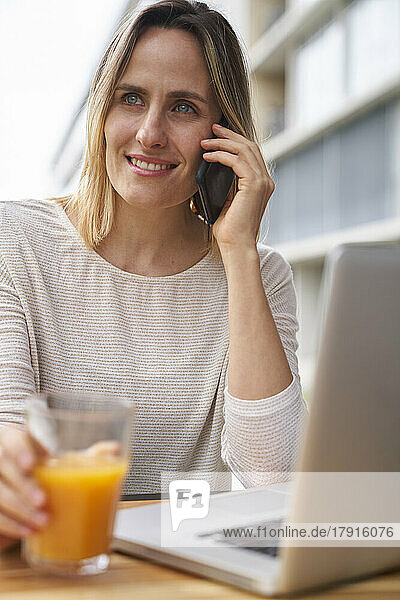 Front view medium shot of woman drinking orange juice in front of laptop computer and speaking on cell phone