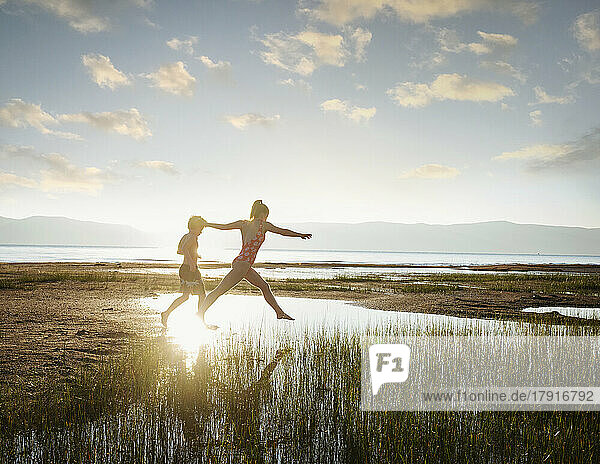 Brother (10-11) and sister (12-13) jumping on stepping stones in lake at sunrise