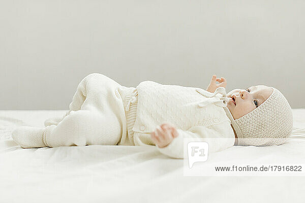 Baby boy (2-5 months) in white outfit lying on bed