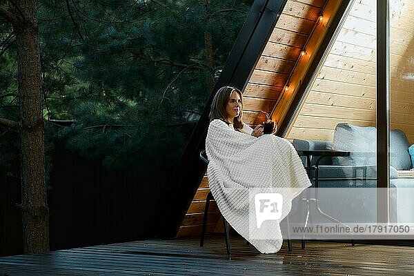 Fröhliche  in eine Decke gehüllte junge Frau sitzt mit einer Tasse heißer Schokolade auf der Terrasse eines Bungalows am Abend