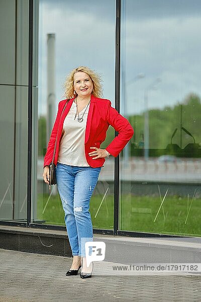 Selbstbewusste Frau in roter Jacke  Bluse und Jeans  die in der Nähe der verstärkten Glaswand des Gebäudes steht
