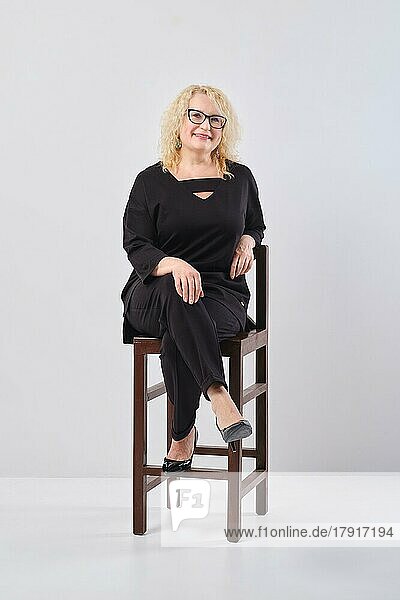 Frau mittleren Alters in schwarzem Outfit und Brille auf einem Stuhl  Studioaufnahme