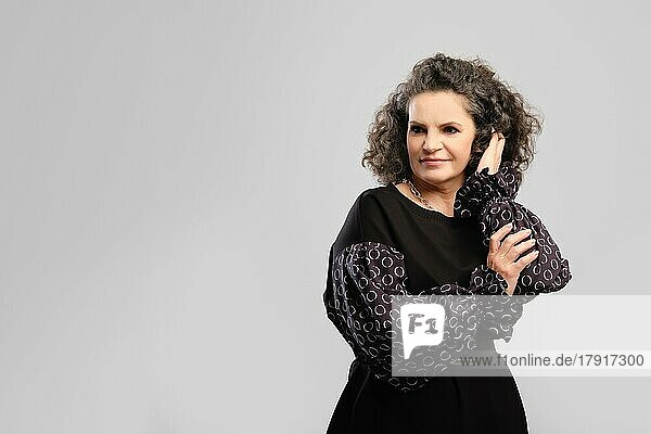 Ältere Frau berührt üppiges lockiges Haar im Studio vor grauem Hintergrund