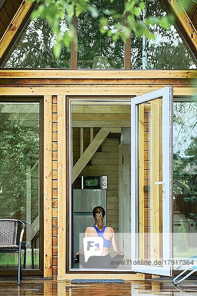 Rückenansicht einer jungen Frau  die in Lotus-Yoga-Pose in einer Holzhütte vor einer offenen Tür sitzt