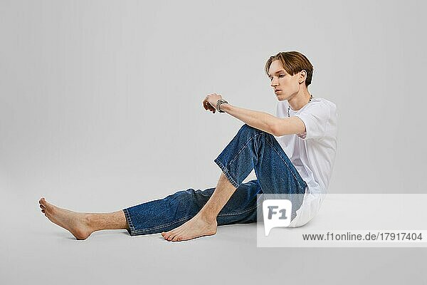 Junger barfüßiger Mann sitzt in T-Shirt und Jeans auf dem Boden im Studio