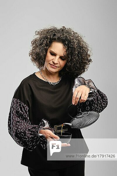 Senior Frau mit grauen Haaren gießt Tee in der Tasse über grauen Studio Hintergrund