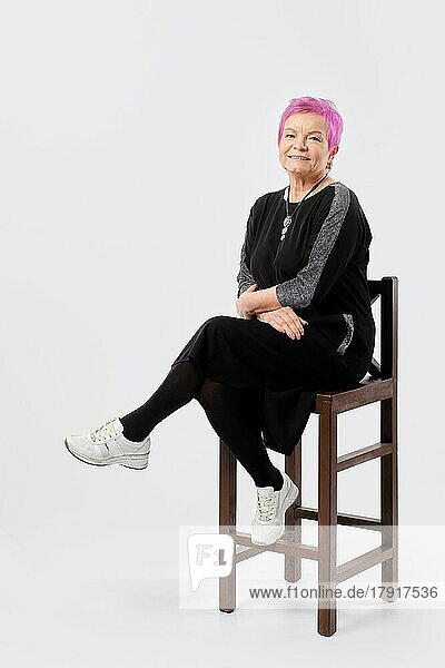 Old woman with short pink hair smiles gently sits on tall chair and keeps hands crossed. Positive emotion concept