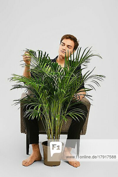Barfüßiger junger Mann in schwarzem Anzug  der in einem Sessel sitzt und eine Zimmerpflanze im Studio betrachtet
