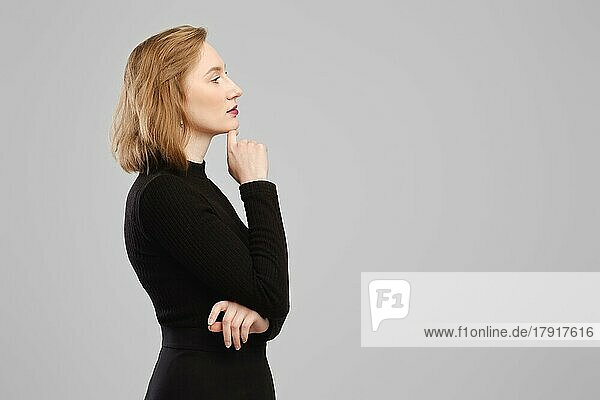 Thoughtful woman standing in profile in studio over grey wall propping up chin and thinking