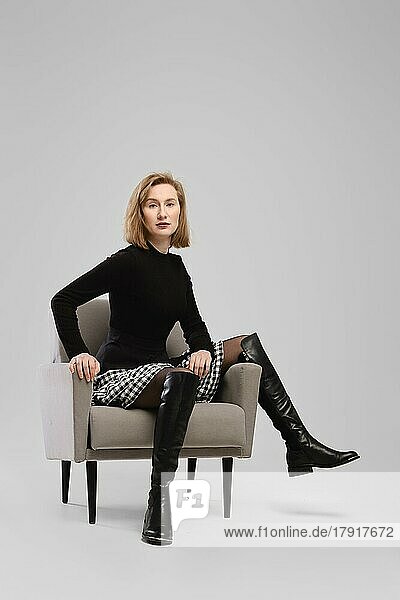 Trendige Frau in Rollkragenpullover und Strapsgürtel über kleinem Rock sitzt in Sessel in Studio über grauem Hintergrund