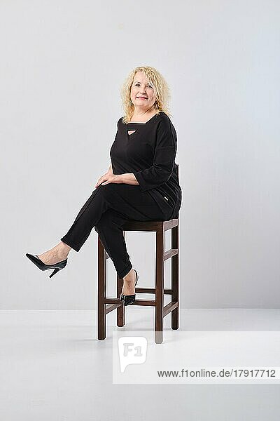 Middle age woman in black outfit sits on chair in white studio