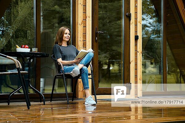 Glückliche Frau sitzt im Korbstuhl auf der Holzterrasse und liest ein Buch
