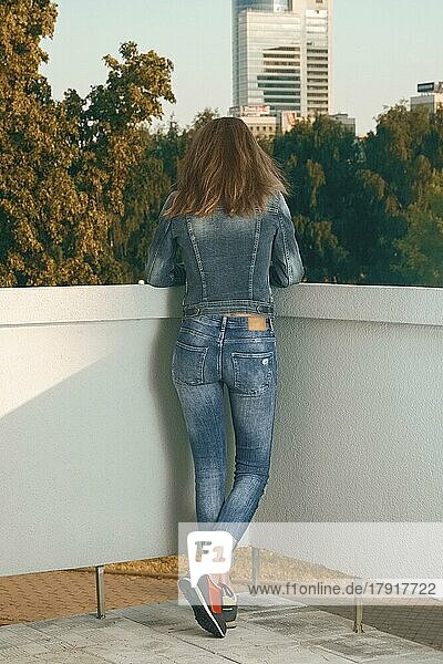 Rückansicht eines schlanken Mädchens in Jeans  das auf einem Balkon steht und nach unten schaut