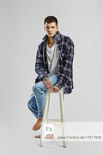 Porträt eines jungen  übermütigen Mannes in Hemd und Jeans  der auf einem hohen Holzstuhl sitzt  in voller Länge