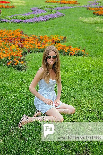 Hübsches Mädchen in kurzen blauen Kleid auf Gras im Sommer Park zwischen Blumen