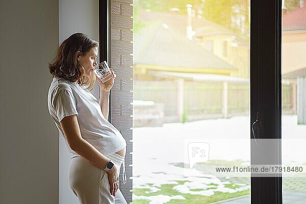 Cute pregnant woman drinks pure water standing by the window in sunny day looking at coming spring