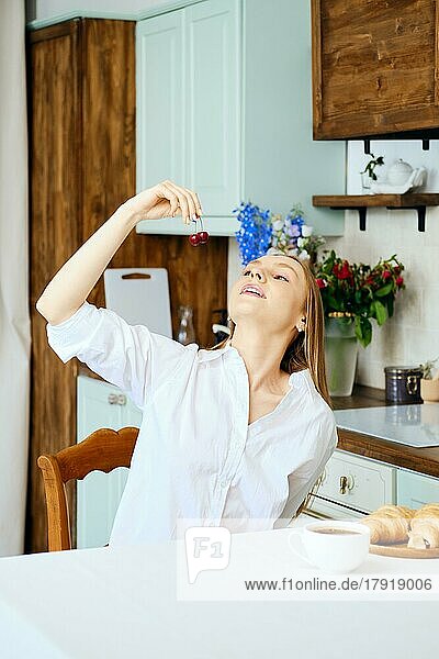 Young woman in white shirt eats sweet cherry in the kitchen