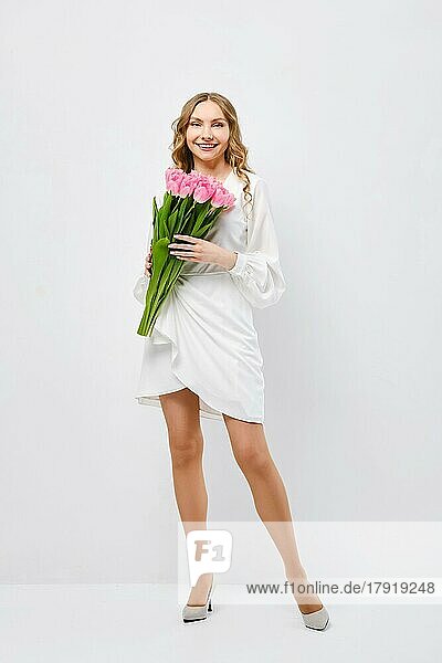 Nette junge fröhliche Frau in weißem Kleid posiert mit Blumenstrauß von Tulpen über weißem Hintergrund