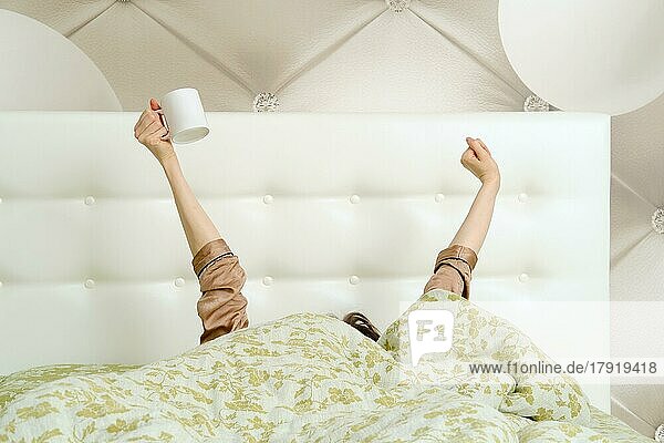 Junge Frau wacht morgens auf  versteckt sich unter der Bettdecke und streckt die Arme mit einer Tasse aus