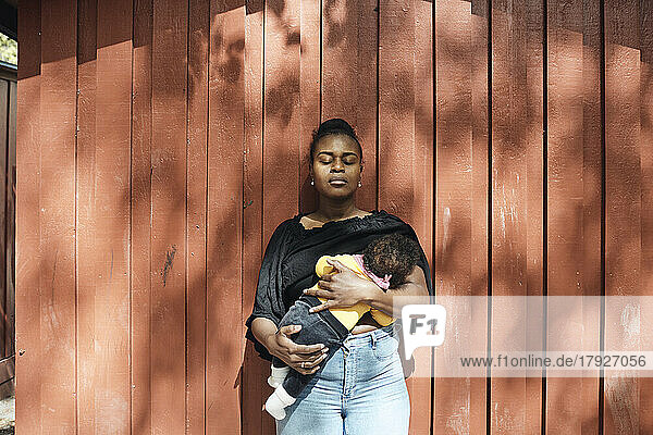 Frau mit geschlossenen Augen stillt ihre kleine Tochter an einem sonnigen Tag vor einer Mauer