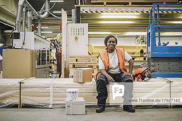 Full length portrait of smiling female carpenter sitting by hardhat in warehouse