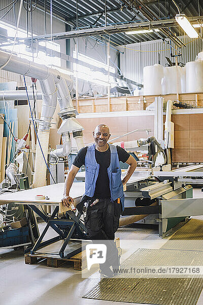 Lächelnder männlicher Zimmermann in voller Länge  der mit der Hand auf der Hüfte an einer Werkbank steht
