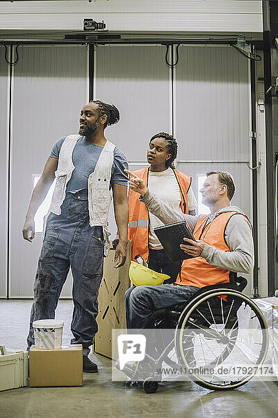 Carpenter sitting on wheelchair with digital tablet instructing male and female coworkers in warehouse