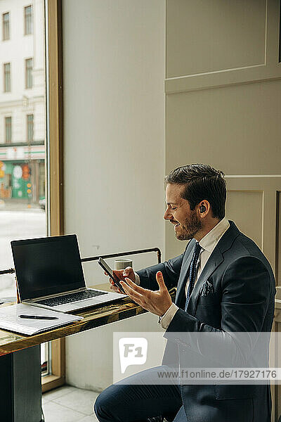 Smiling businessman holding mobile phone while talking through wireless-in ear headphones at office