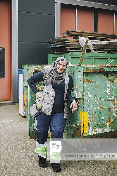 Ganzkörperporträt eines lächelnden weiblichen Bauarbeiters  der mit der Hand auf der Hüfte neben einem Metallbehälter steht