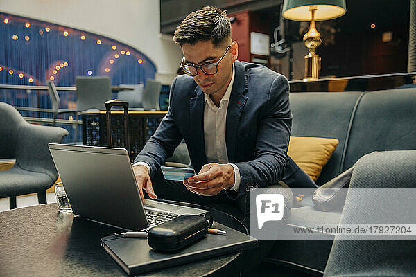 Businessman holding credit card while using laptop sitting in hotel lounge
