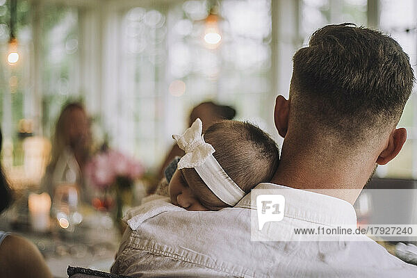 Tochter mit Stirnband schläft auf der Schulter des Vaters bei einer Dinnerparty