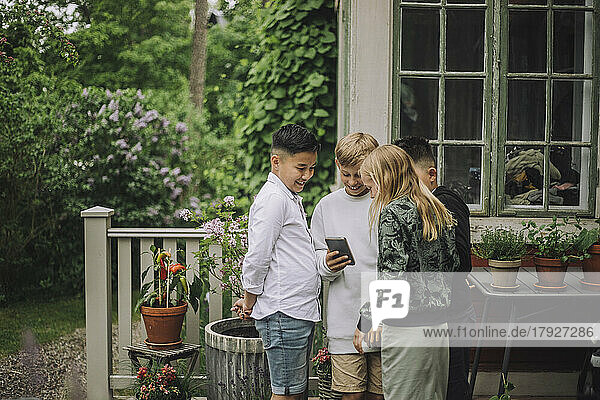 Lächelnde männliche und weibliche Freunde teilen sich ein Mobiltelefon  während sie in der Nähe eines Hauses stehen