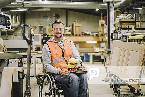Portrait of mature carpenter with ear protectors and digital tablet sitting on wheelchair in warehouse