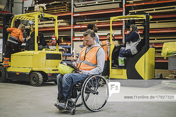 Männlicher Tischler mit Behinderung  der im Rollstuhl sitzend ein digitales Tablet benutzt  in voller Länge im Lager