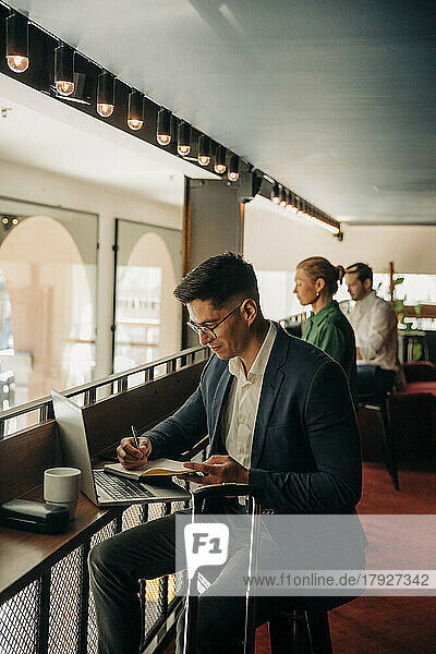 Geschäftsmann schreibt in Tagebuch  während er mit Laptop im Hotel sitzt