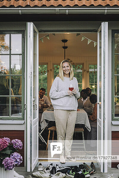 Porträt einer lächelnden reifen Frau mit Getränk an der Tür stehend
