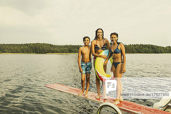 Lächelndes Mädchen mit aufblasbarem Ring steht mit Geschwistern auf einem Sprungbrett am See